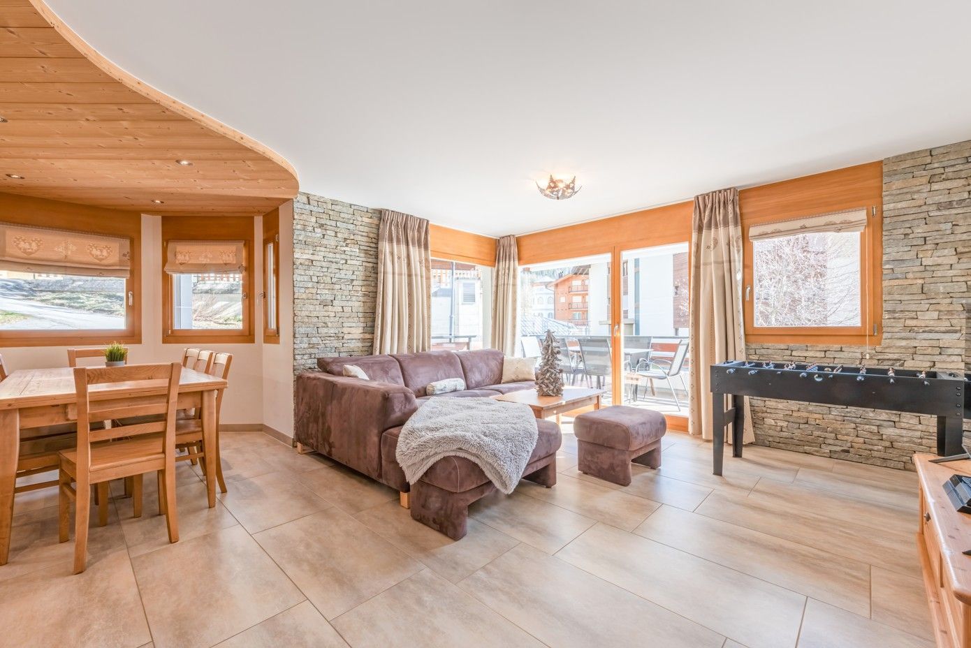 Salon avec plafond en bois, mur en pierre, canapé, table à manger et baby-foot.