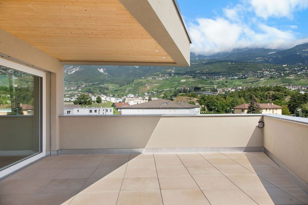 Balcon vide au sol clair et au mur beige, donnant sur une ville et des montagnes sous un ciel bleu.