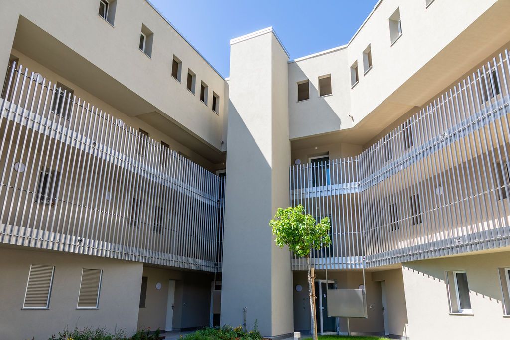 Bâtiment moderne aux murs gris, aux claustras et avec un petit arbre dans une cour.