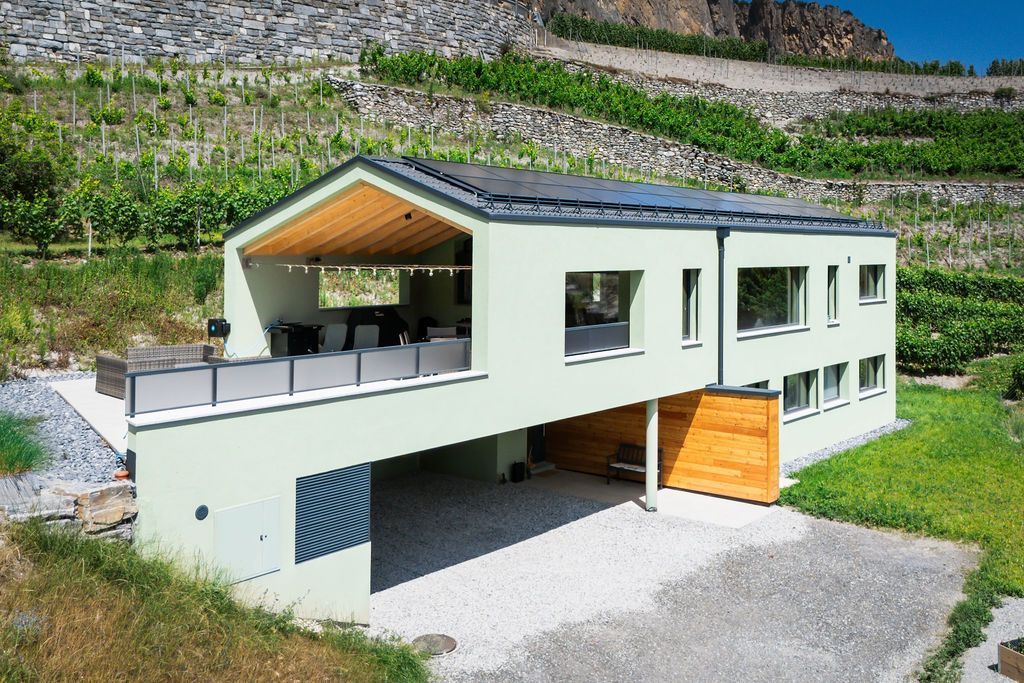 Maison moderne de deux étages avec une façade vert clair, des éléments en bois et un balcon donnant sur une colline couverte de vignes.