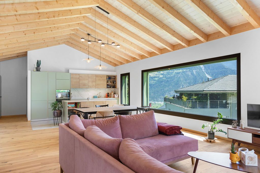Intérieur avec plafond en bois, canapé rose, cuisine et grande fenêtre donnant sur la montagne.