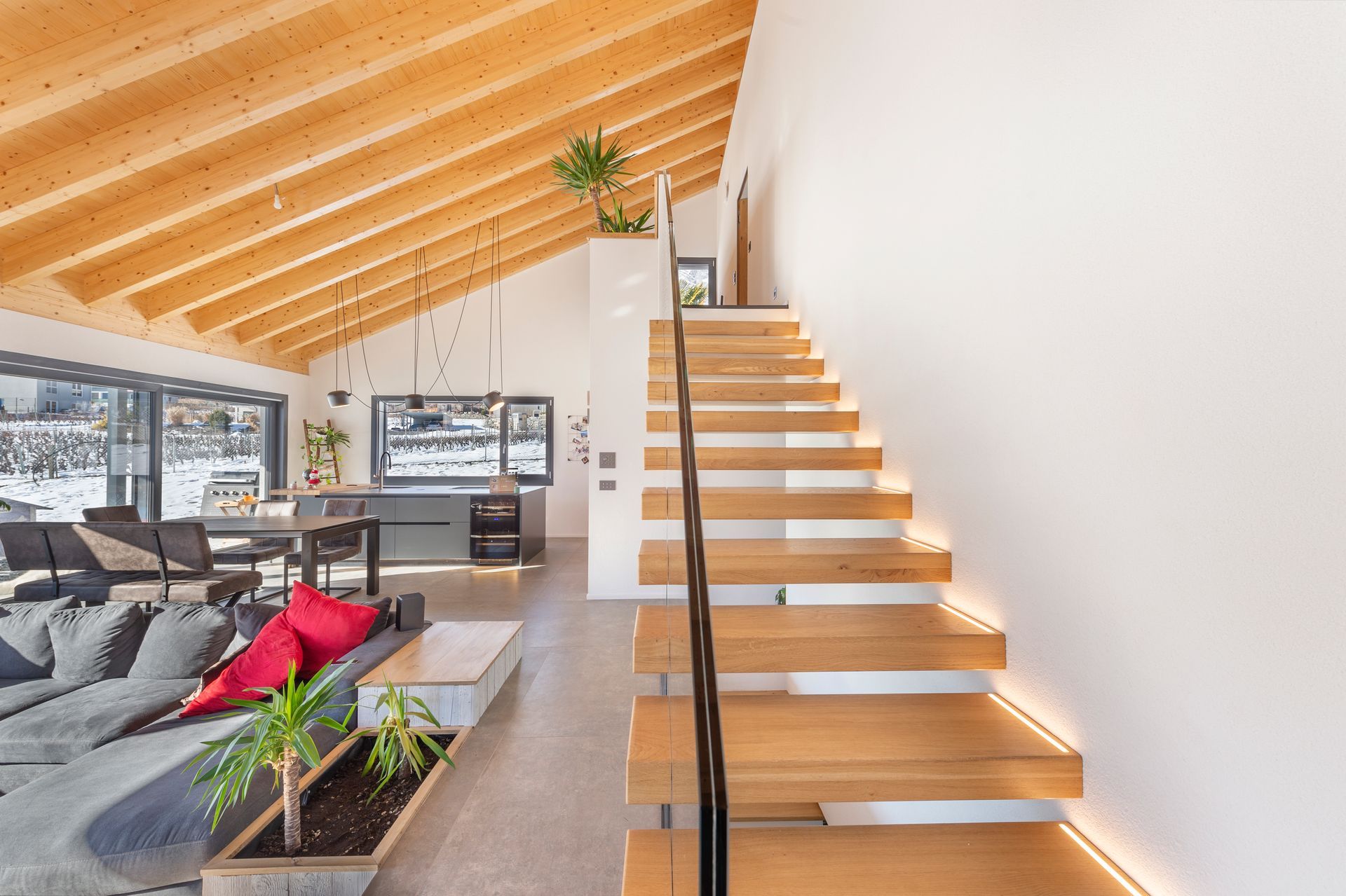 Espace de vie à aire ouverte avec escalier en bois, plafond et grandes fenêtres donnant sur un paysage enneigé.