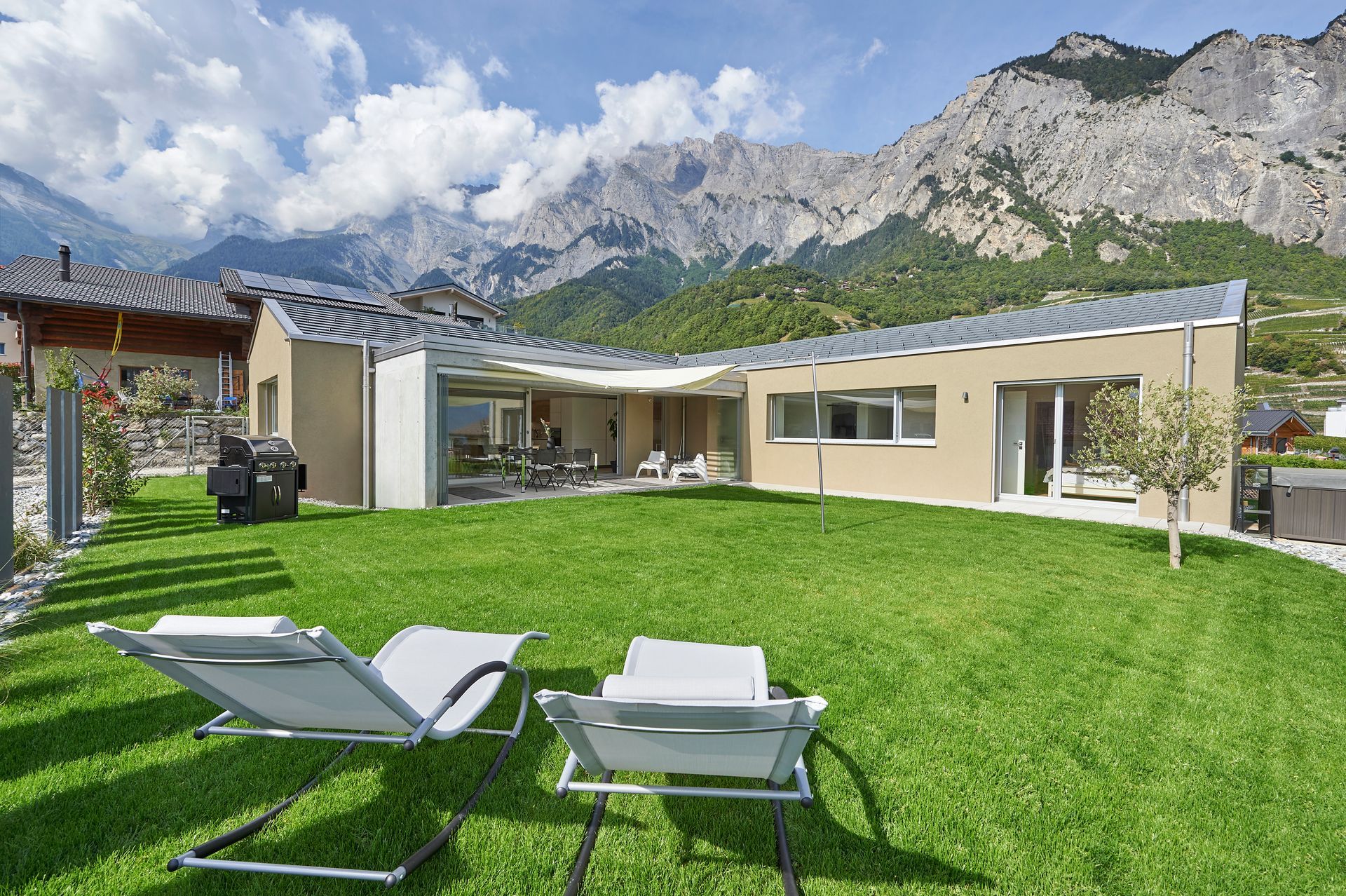 Maison moderne avec pelouse et vue sur la montagne, deux chaises longues sur l'herbe verte.