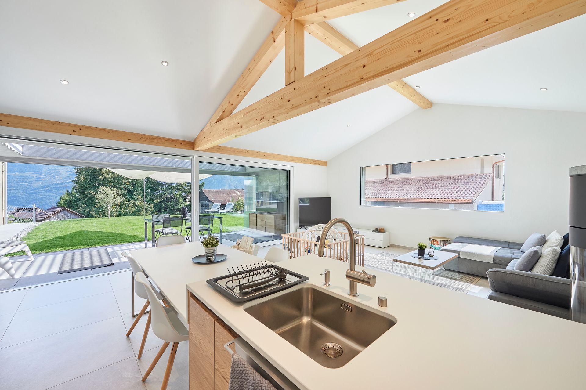 Cuisine moderne avec îlot central, poutres apparentes et vue dégagée sur le jardin et les montagnes.