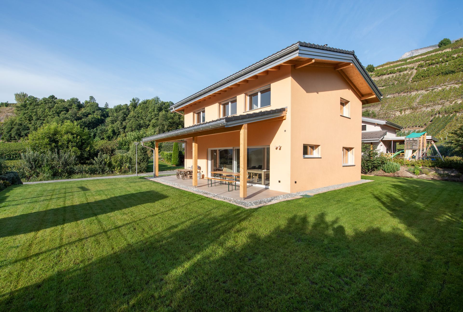Maison à deux étages avec façade couleur pêche, éléments en bois et patio couvert, située dans un jardin verdoyant avec un vignoble en arrière-plan.