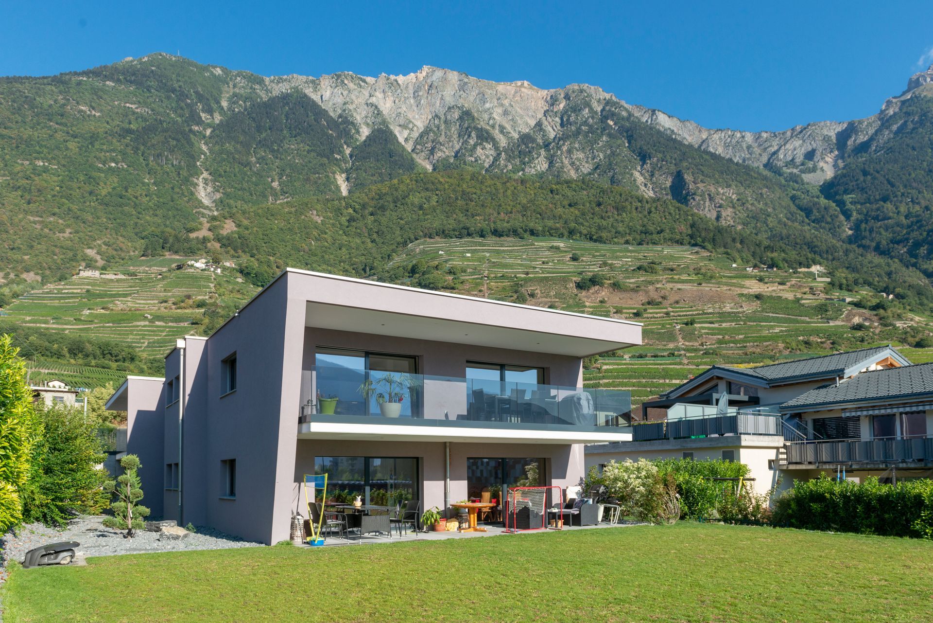Maison moderne avec balcon et pelouse, avec une chaîne de montagnes en toile de fond, sous un ciel bleu azur.