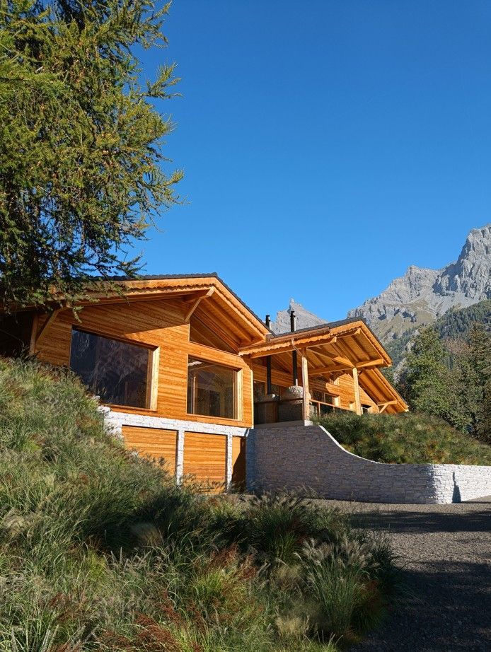 Cabane en bois nichée à flanc de colline, avec les montagnes en toile de fond, sous un ciel d'un bleu limpide.