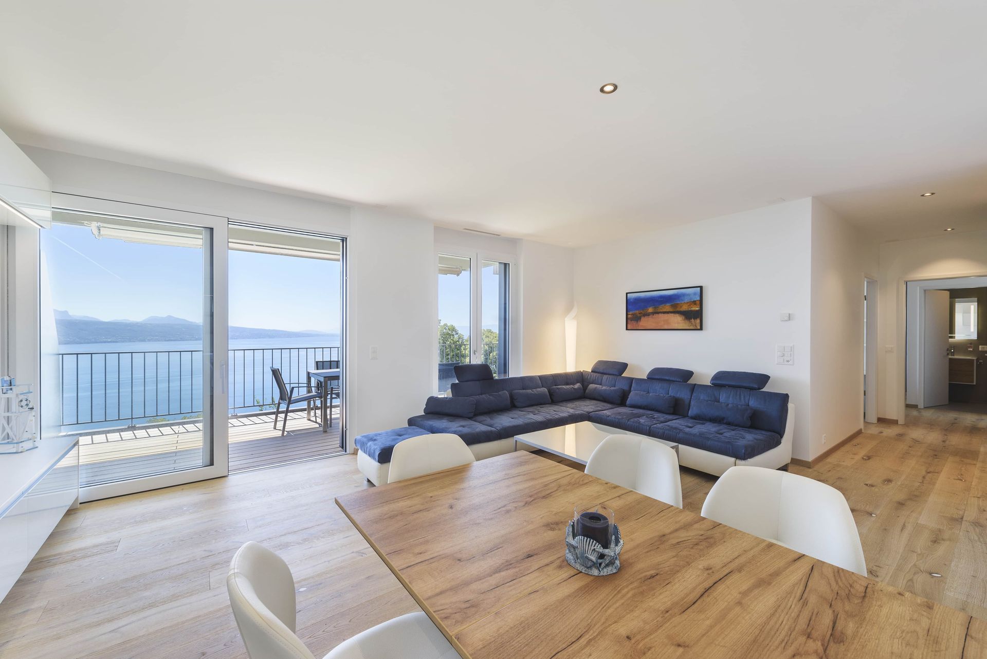 Salon lumineux avec vue sur l'eau, canapé d'angle bleu, table à manger et accès au balcon.