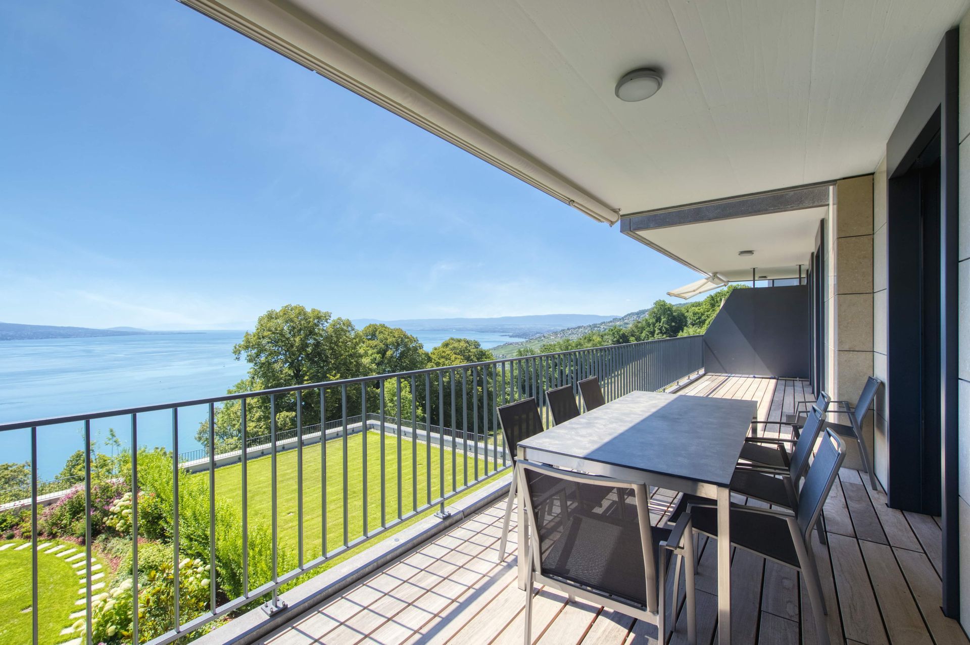Balcon donnant sur un lac, avec table à manger, chaises et rambarde en métal. Pelouse verdoyante en contrebas. 