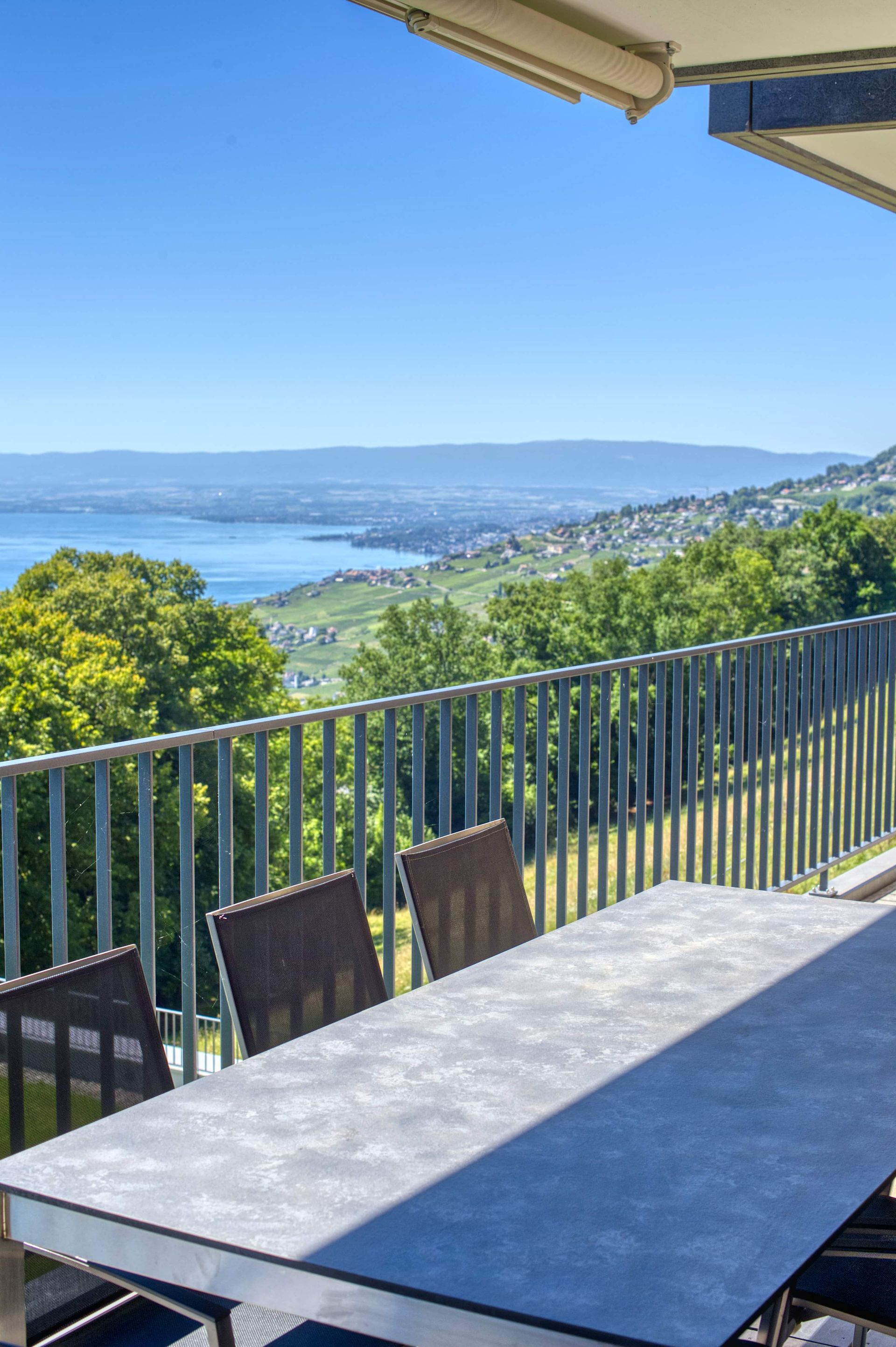 Table à manger extérieure avec chaises sur un balcon donnant sur un lac et une colline au loin, sous un ciel bleu azur.