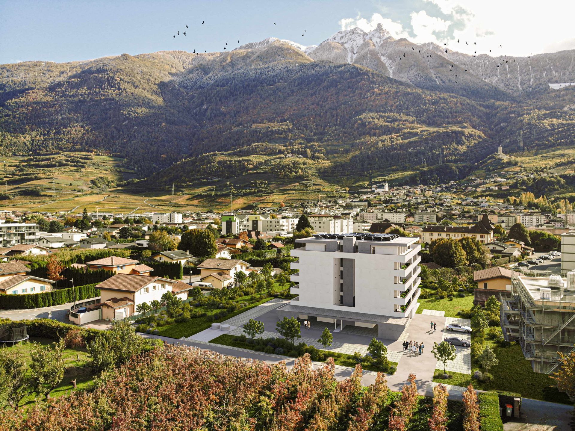 Bâtiment blanc moderne dans une vallée verdoyante, avec des montagnes en arrière-plan. Voitures garées devant.