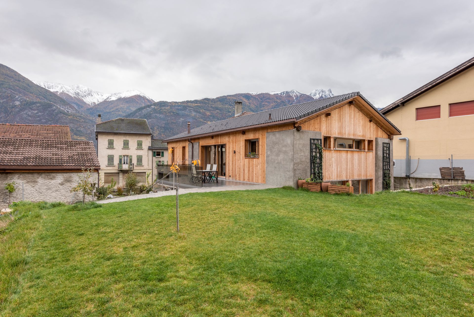 Une maison en bois avec une pelouse verte, des montagnes en arrière-plan, un ciel nuageux.