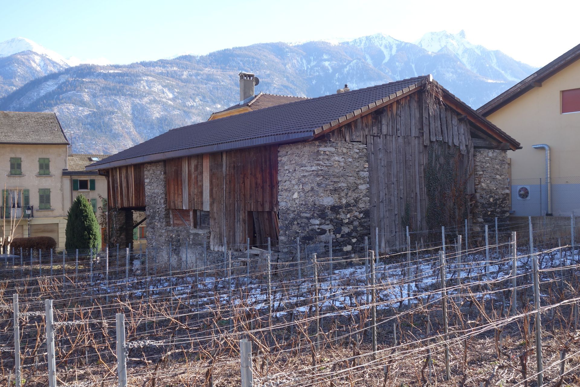 Vieille grange en bois avec soubassement en pierre, vignes et montagnes enneigées en arrière-plan.