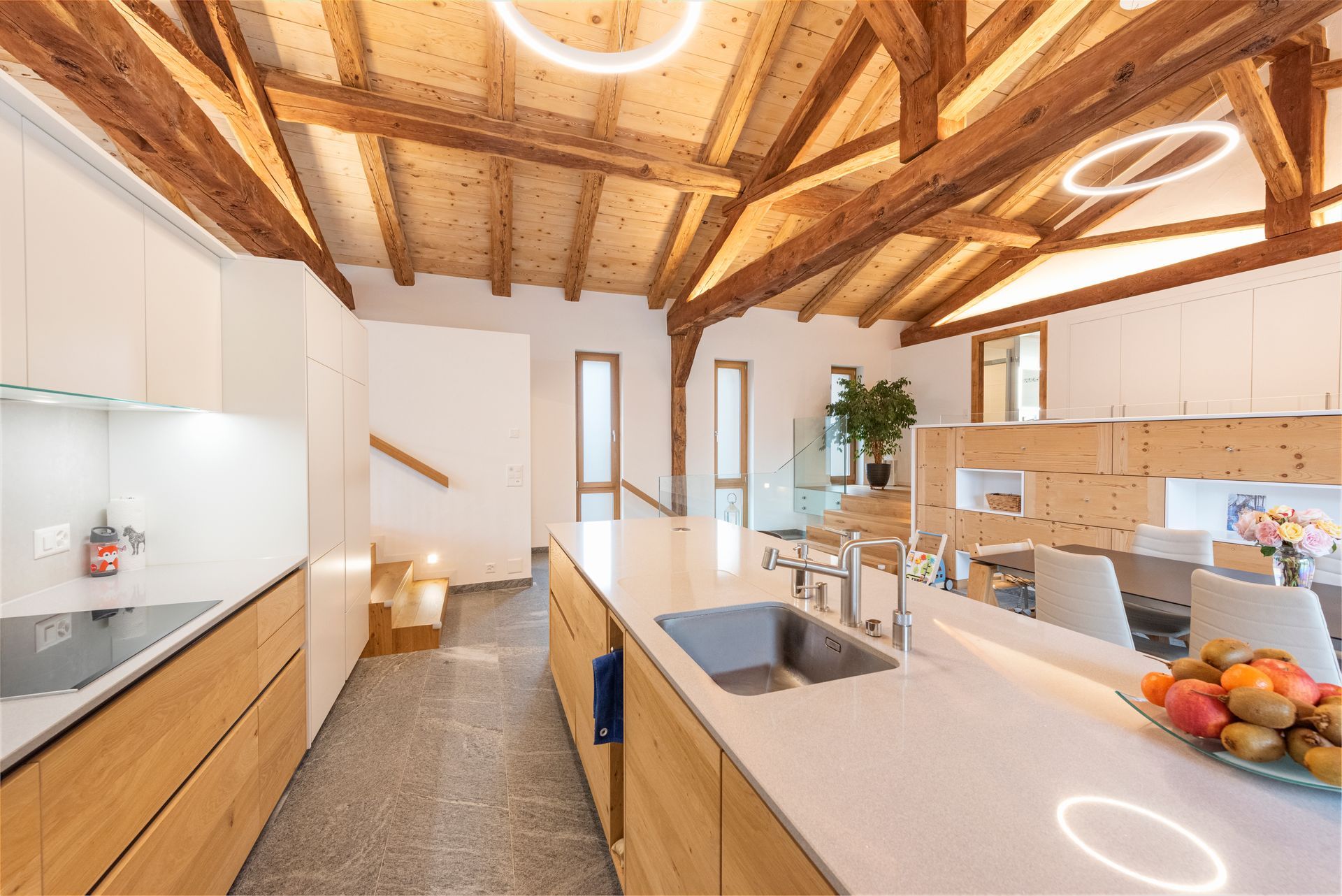 Cuisine moderne avec poutres apparentes, placards blancs, îlot central avec évier et corbeille à fruits.