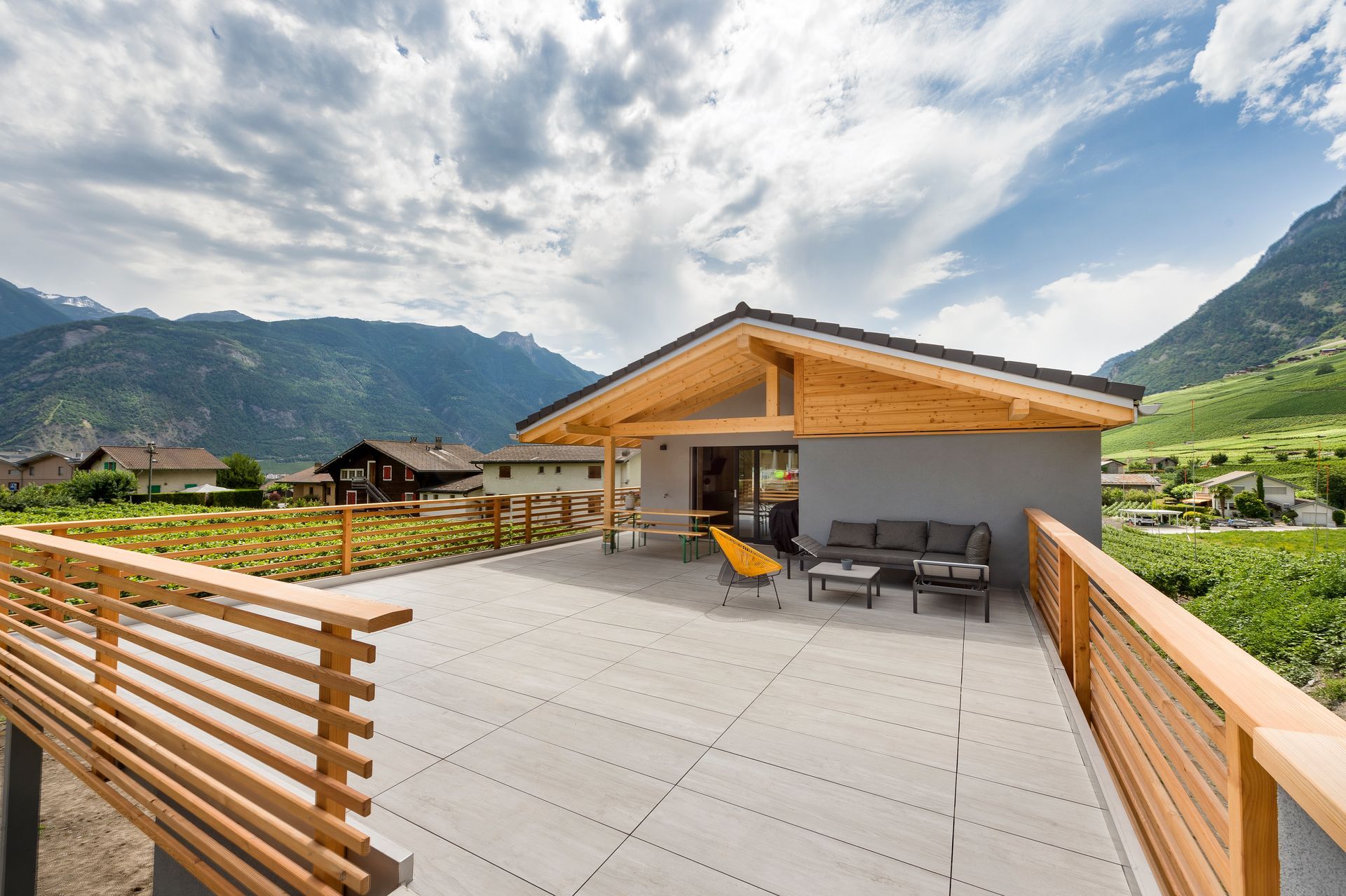 Maison moderne avec une grande terrasse et vue sur la montagne. Poutres apparentes, murs gris et coin salon extérieur.