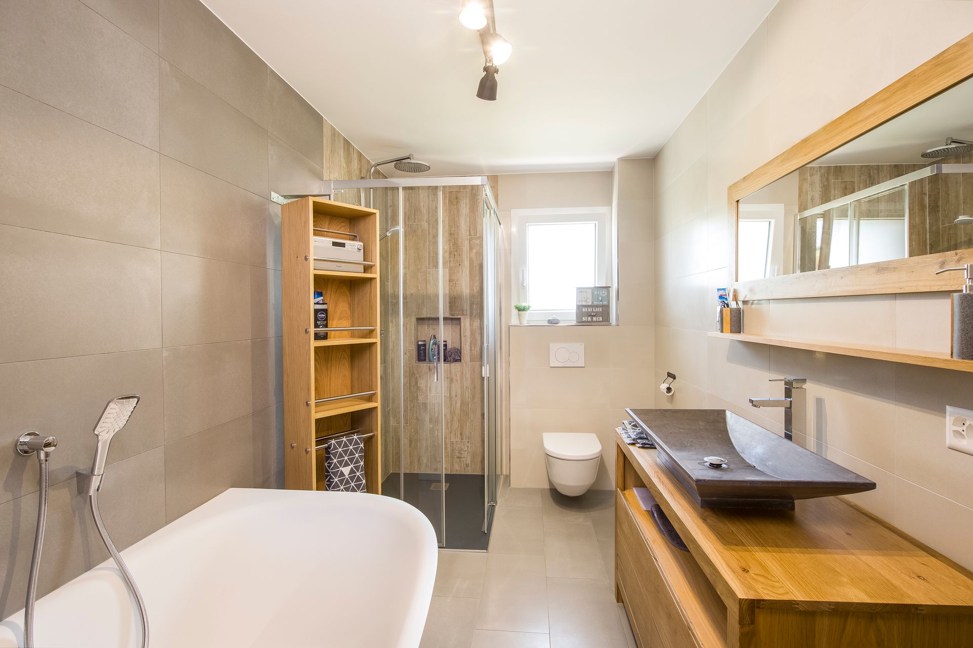 Salle de bains moderne avec baignoire, douche, meuble-lavabo en bois et toilettes. Carrelage beige et lumière naturelle.