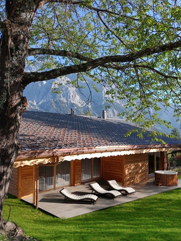 Chalet en bois avec terrasse, chaises longues et jacuzzi. Montagnes en arrière-plan.