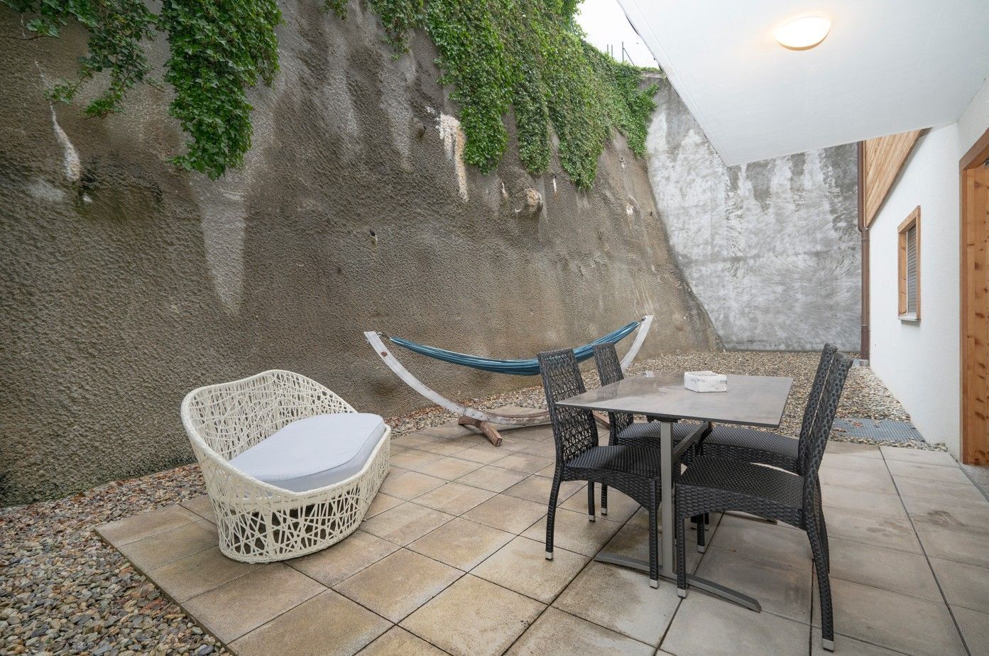 Terrasse avec hamac, table et chaises, adossée à un mur de pierre recouvert de vigne.