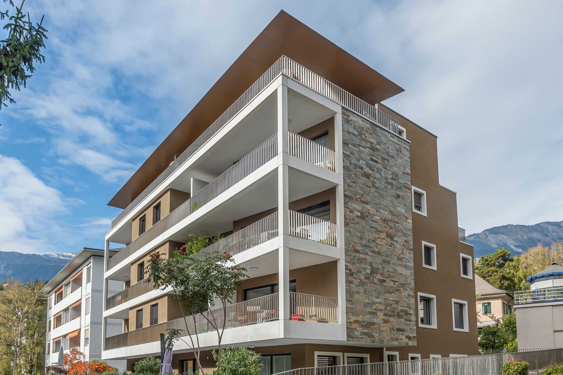 Immeuble d'appartements moderne avec balcons, façade marron, mur en pierre, ciel bleu.