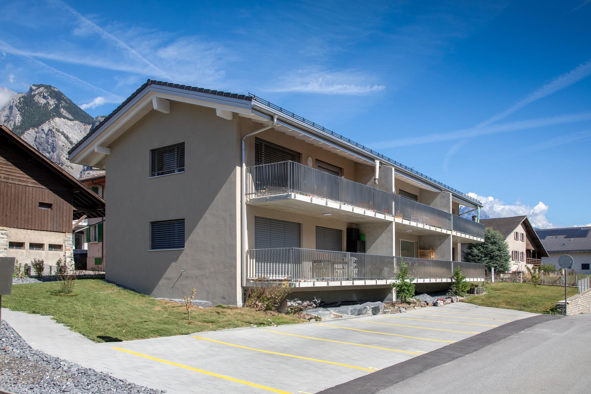 Immeuble d'appartements avec balcons, parking en gravier et vue sur les montagnes sous un ciel bleu.