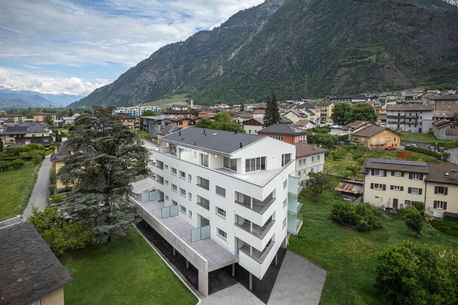 Immeuble d'appartements blanc moderne en ville, avec des montagnes en arrière-plan. Environnement verdoyant.