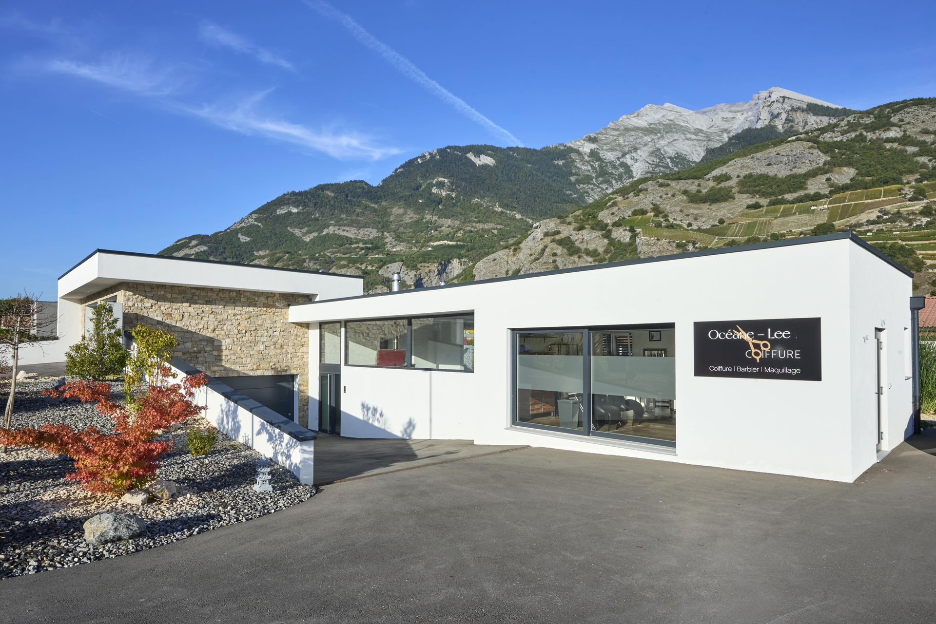 Bâtiment blanc moderne avec de grandes fenêtres et une enseigne, montagnes en arrière-plan.