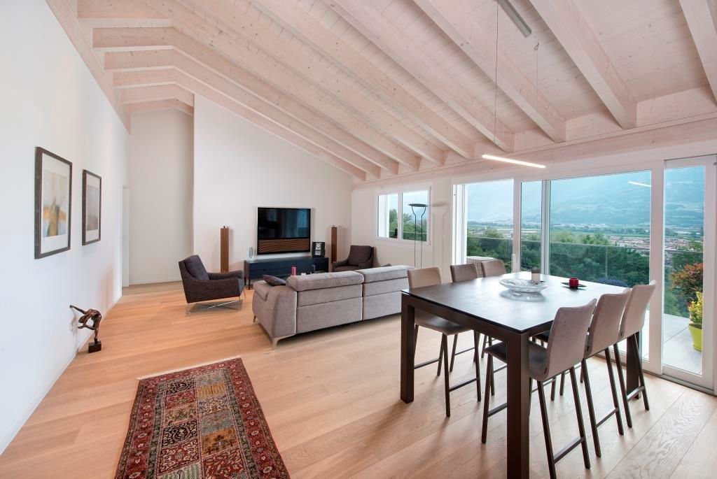 Espace de vie spacieux avec poutres apparentes, parquet clair, tapis, table à manger et grandes fenêtres offrant une vue.