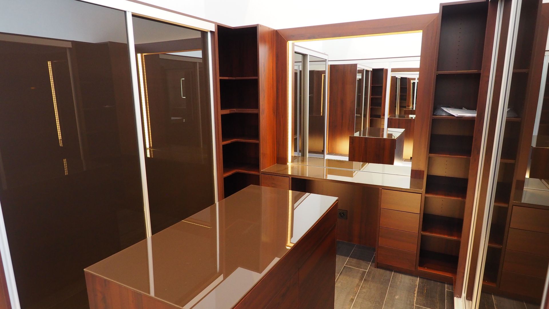 Dressing moderne avec étagères en bois foncé, tiroirs et portes vitrées. Les murs en miroir reflètent l'espace.