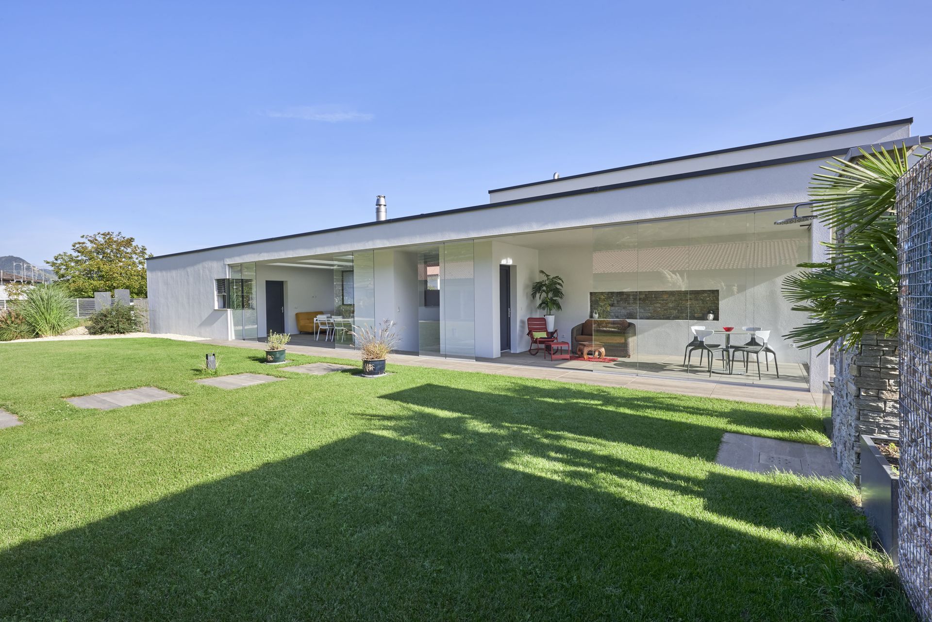 Vue extérieure d'une maison blanche moderne avec pelouse verte et terrasse par une journée ensoleillée.