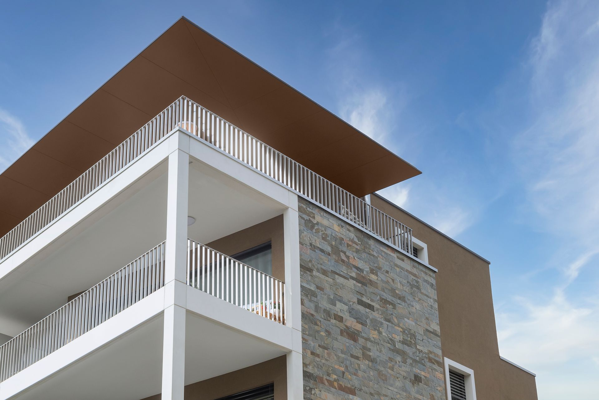 Immeuble moderne avec balcons blancs, façade en pierre, toit brun, se détachant sur un ciel bleu.