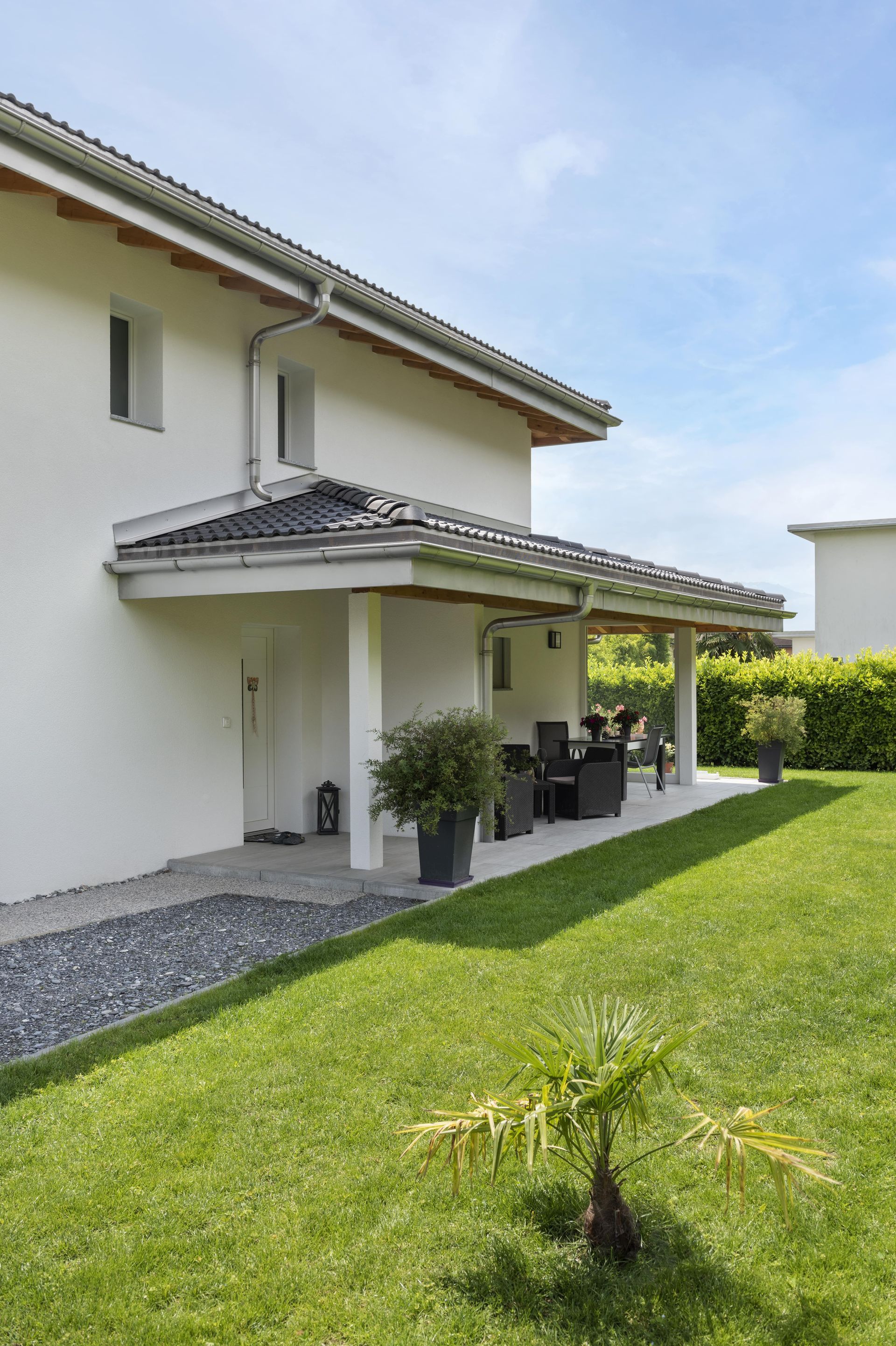 Maison blanche avec patio couvert, aménagé avec du mobilier de jardin et des plantes en pot sur une pelouse verte.
