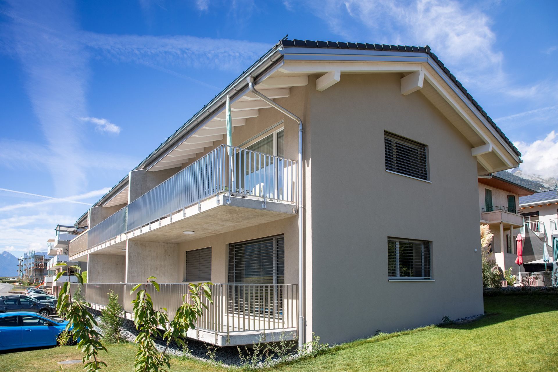 Immeuble beige de deux étages avec balcons. Ciel bleu, voitures garées à proximité.