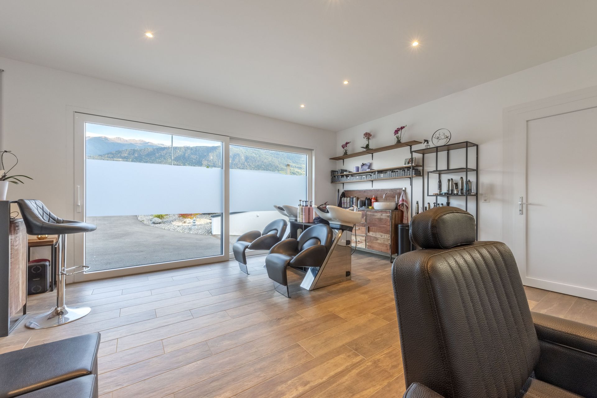 Intérieur d'un salon de coiffure avec fauteuils, lavabo et étagères. Grande fenêtre en arrière-plan.