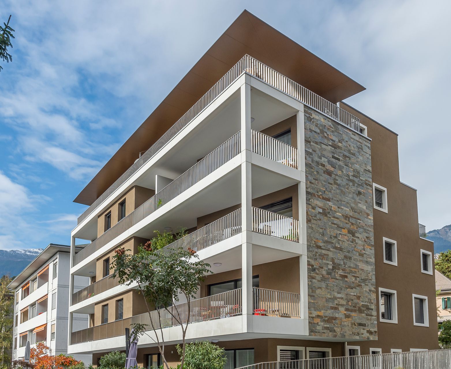Immeuble d'appartements moderne avec façade en stuc brun et en pierre, balcons et toit plat sur fond de ciel bleu.