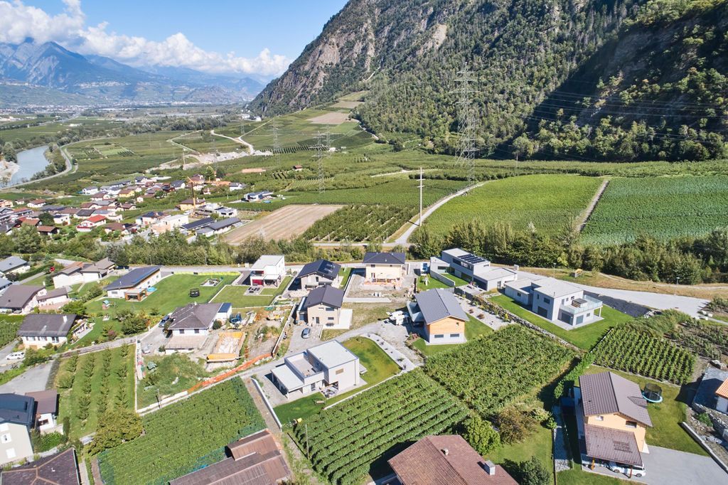 Vue aérienne d'une vallée avec des maisons, des champs, des montagnes et une rivière. Les teintes vertes et brunes dominent le paysage.