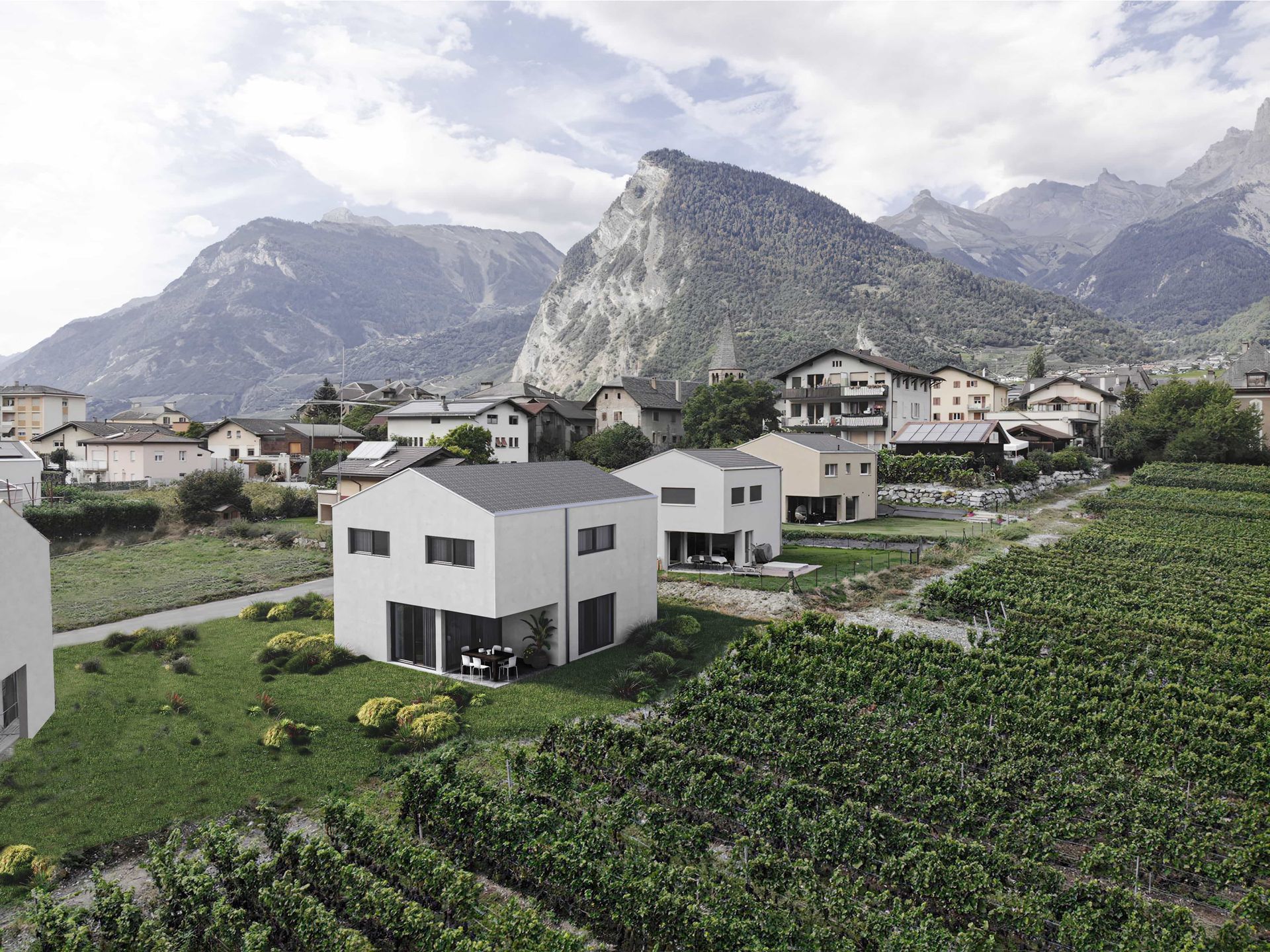 Maisons blanches modernes dans une vallée entourée de vignobles et de montagnes, sous un ciel nuageux.