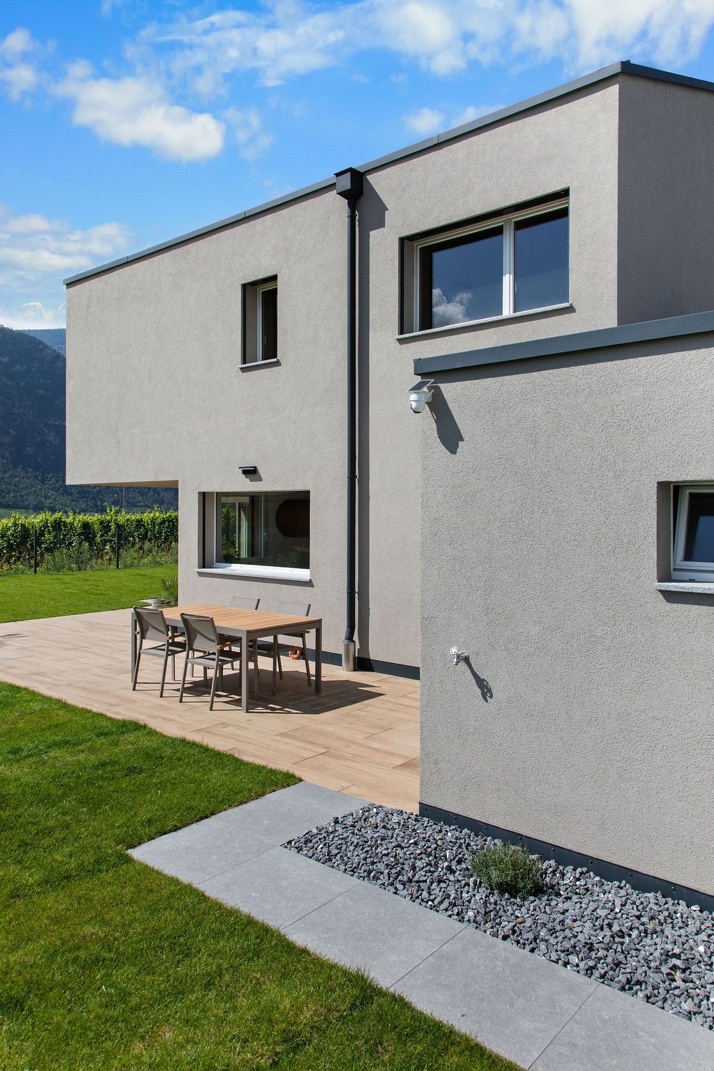 Maison grise moderne avec coin salon extérieur sur une terrasse en bois, pelouse verte et ciel bleu.