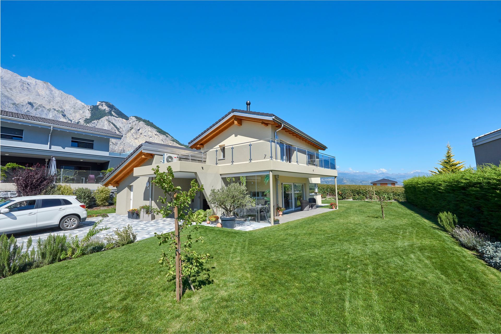 Maison à deux étages avec pelouse verte, voiture, montagnes en arrière-plan sous un ciel bleu.