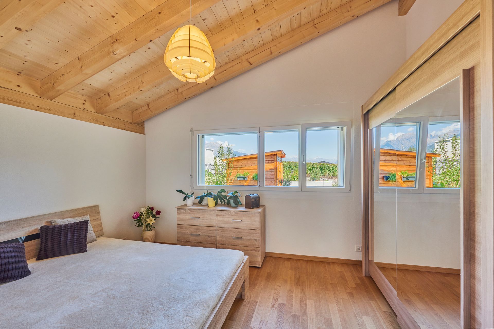Chambre avec plafond et parquet en bois, lit, commode, grande fenêtre et portes de placard à miroir.