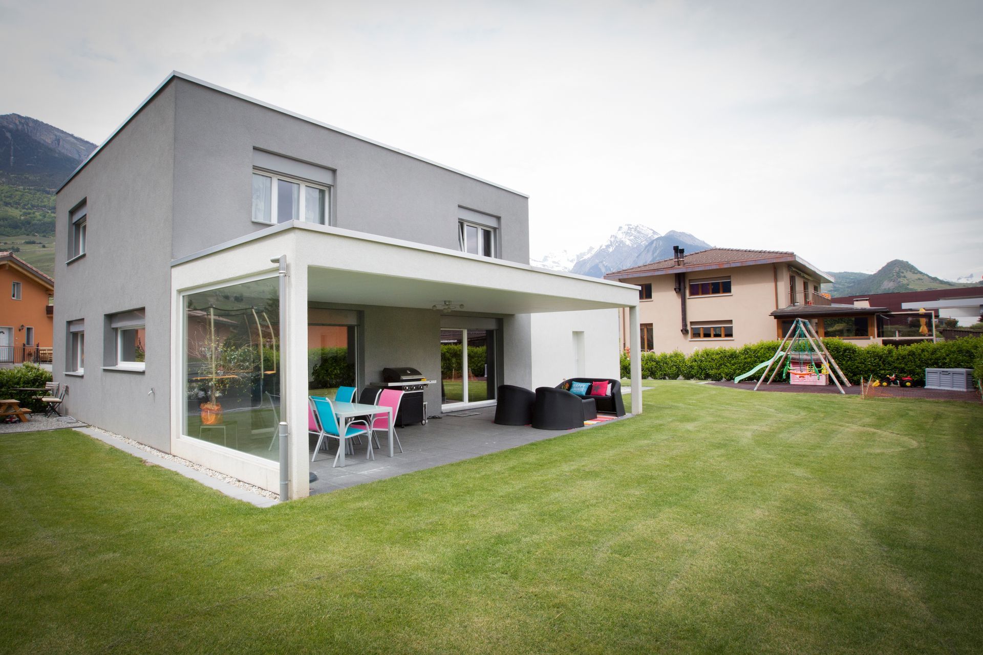 Maison grise moderne avec terrasse, mobilier de jardin et jardin gazonné avec vue sur les montagnes.