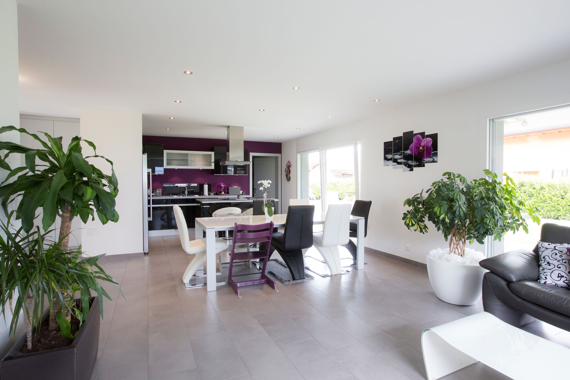 Espace salle à manger et cuisine ouverte avec table et chaises blanches, cuisine noire et violette et plantes en pot.