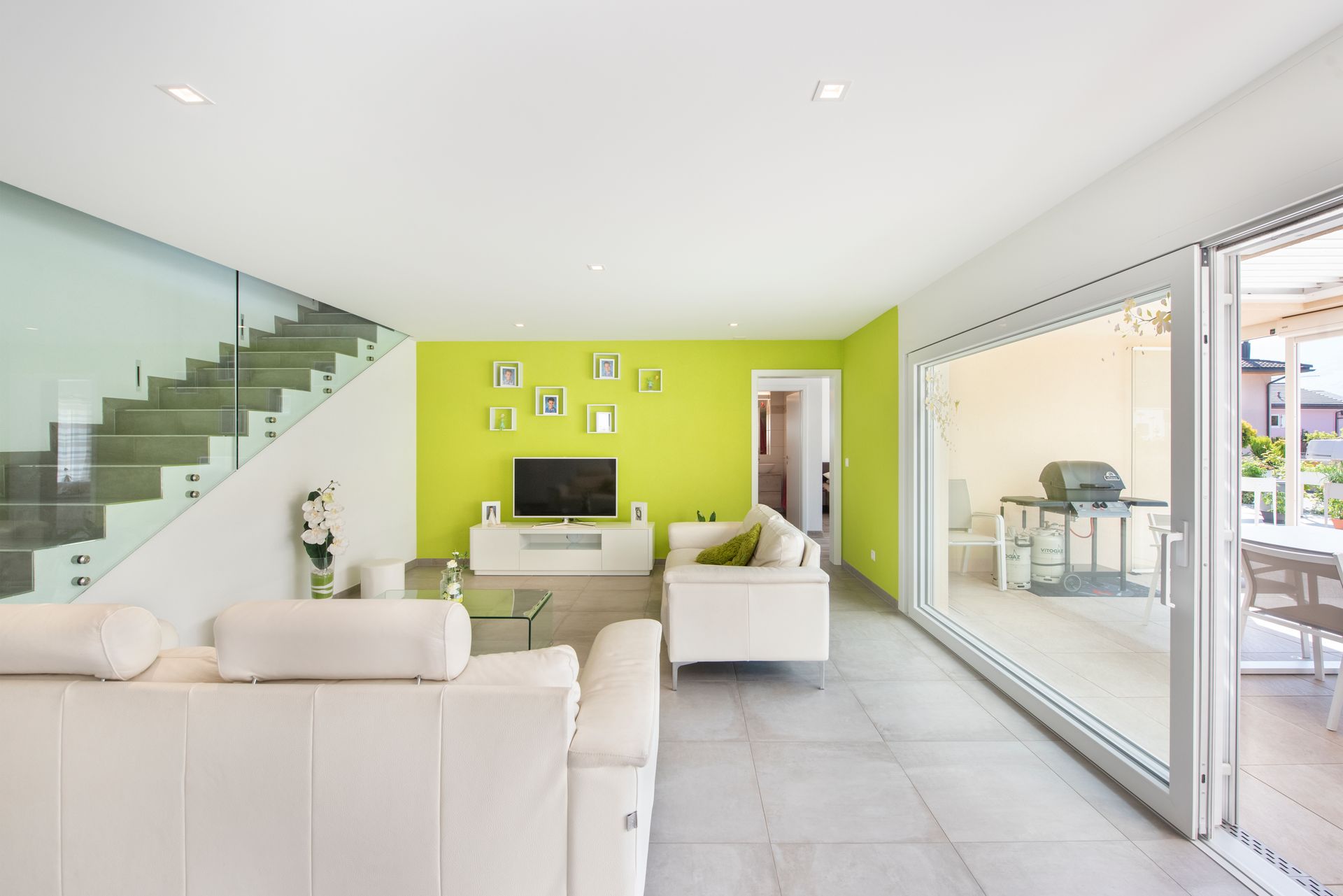 Salon lumineux avec meubles blancs, murs vert citron, escalier en verre et porte coulissante donnant sur le patio.