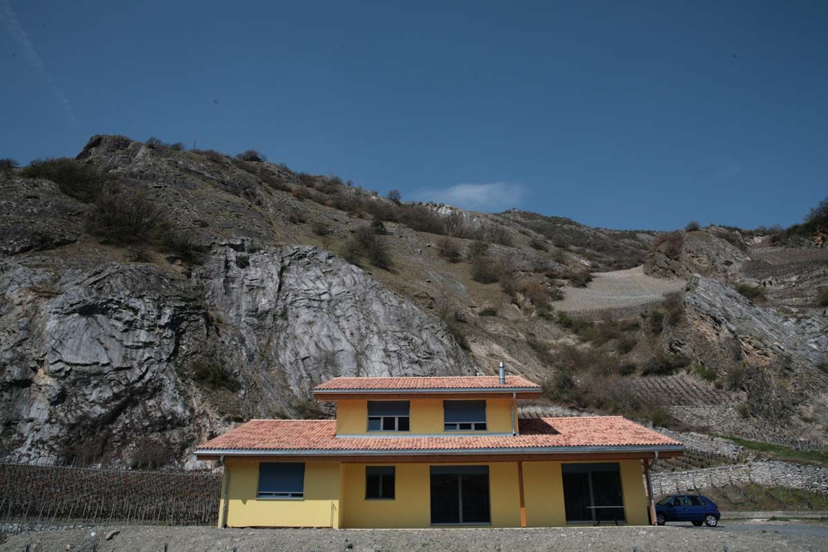 Maison jaune au toit orange devant un flanc de montagne rocheux sous un ciel bleu.