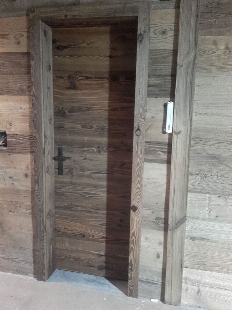 Eine Holztür, eingelassen in eine rustikale, holzgetäfelte Wand, mit einem weißen, an der Wand montierten Schalter oder Sensor in der Nähe.