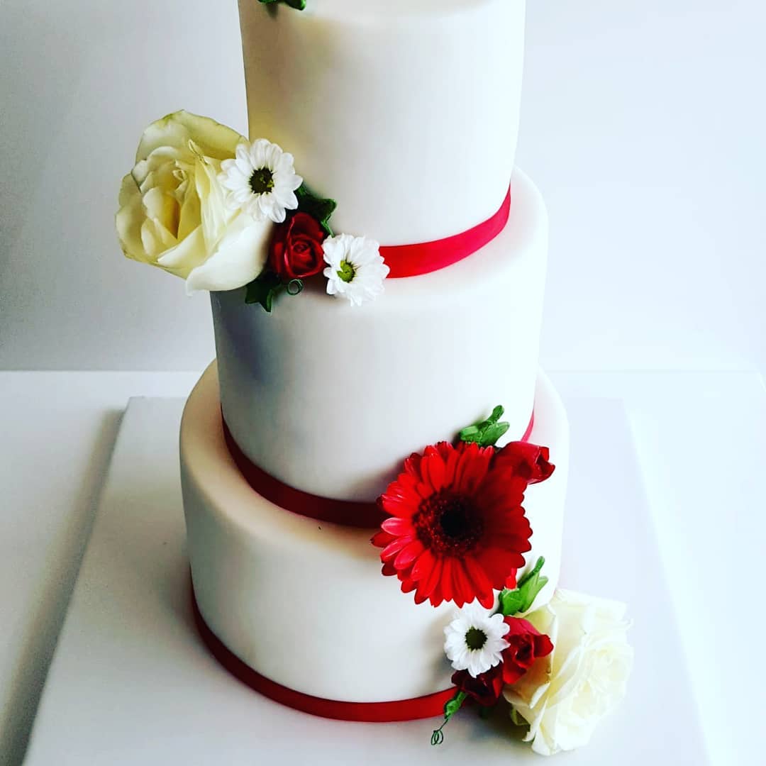 Gâteau blanc à trois étages avec ruban rouge et décorations florales, notamment des gerberas rouges et des roses blanches.