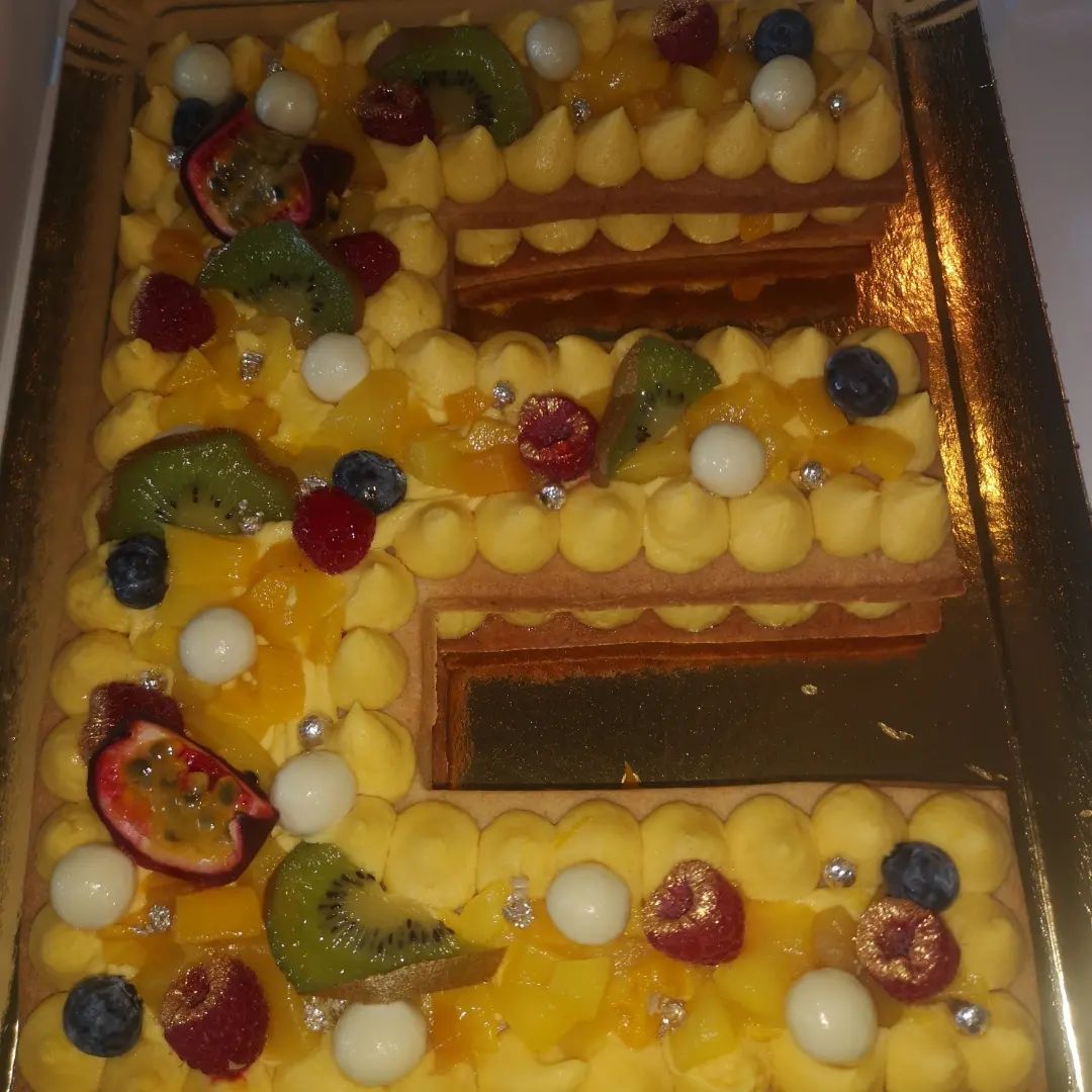 Dessert pâtissier en forme de lettre avec glaçage jaune, fruits et boules de chocolat blanc.