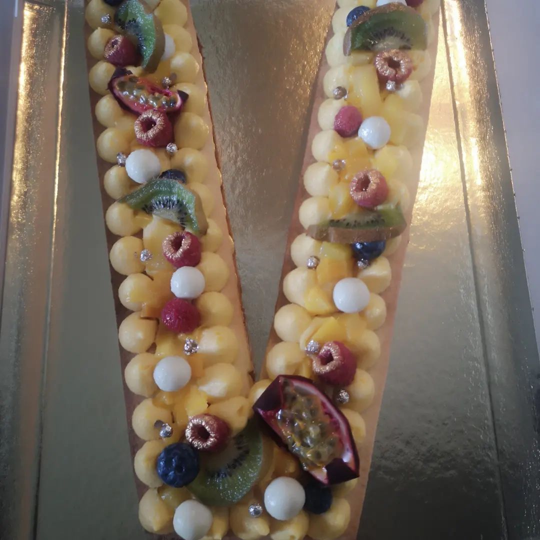 Gâteau en forme de lettre « V » avec glaçage jaune, fruits frais et bonbons blancs sur un plateau doré.