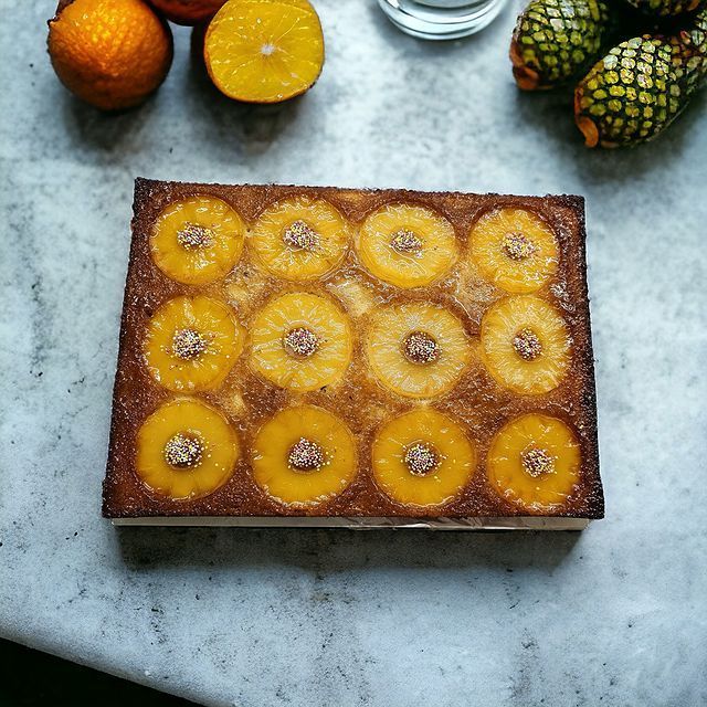 Gâteau renversé à l'ananas avec des tranches d'orange et des fruits verts dispersés sur une surface en marbre blanc.