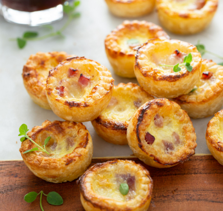 Mini quiches à la croûte dorée, garnies d'oeuf et de jambon, sur une planche de bois, garnies d'herbes.