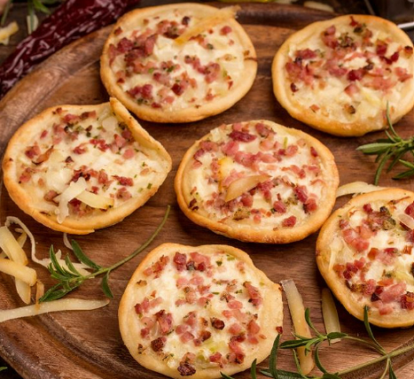 Mini tartelettes garnies de bacon, d'oignons et de fromage disposées sur un plateau en bois avec des herbes et un poivron.